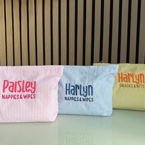 K&ouml;nnte beinhalten: Drei Seersucker-Stoffbeutel in Rosa, Blau und Gelb. Jeder Beutel hat einen anderen Namen aufgestickt: "Paisley", "Harlyn" und "Harlyn". Auf dem rosa Beutel steht "Nappies & Wipes". Auf dem blauen Beutel steht "Nappies & Wipes". Auf dem gelben Beutel steht "Snacks & Bits".