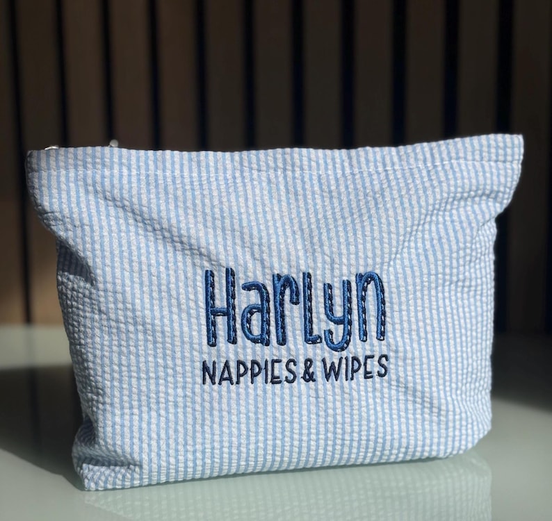May include: A blue and white striped seersucker pouch. The pouch has the name "Harlyn" embroidered in dark blue, with the words "Nappies & Wipes" underneath. Ideal for storing baby nappies and wipes.