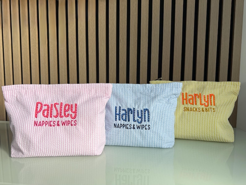 K&ouml;nnte beinhalten: Drei Seersucker-Stoffbeutel in Rosa, Blau und Gelb. Jeder Beutel hat einen anderen Namen aufgestickt: "Paisley", "Harlyn" und "Harlyn". Auf dem rosa Beutel steht "Nappies & Wipes". Auf dem blauen Beutel steht "Nappies & Wipes". Auf dem gelben Beutel steht "Snacks & Bits".