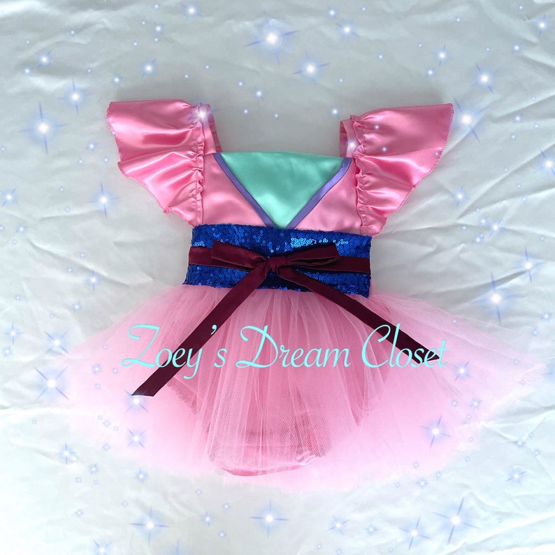 May include: Pink tulle tutu dress with a pink satin bodice, mint green and purple accents, a blue sequin belt, and a burgundy bow. The dress is for a child and has the text "Zoey's Dream Closet" on the tulle skirt.
