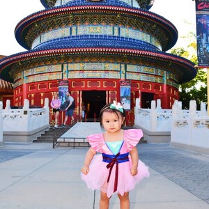 May include: A young girl wearing a pink and blue dress with a red sash stands in front of a traditional Chinese pagoda. The pagoda is decorated with red, blue, and gold accents. The girl is smiling and looking at the camera.