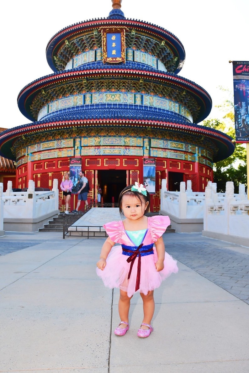 May include: A young girl wearing a pink and blue dress with a red sash stands in front of a traditional Chinese pagoda. The pagoda is decorated with red, blue, and gold accents. The girl is smiling and looking at the camera.
