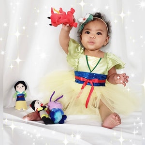 May include: A young girl dressed as Mulan in a yellow and green dress with a blue and red sash. She is holding a red dragon plush toy and is surrounded by other plush toys, including a purple bug and a girl with black hair.
