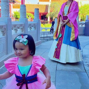 May include: A young girl in a pink and teal dress with a flower in her hair walks towards a woman in a pink and blue dress. The woman is standing in front of a traditional Chinese building.