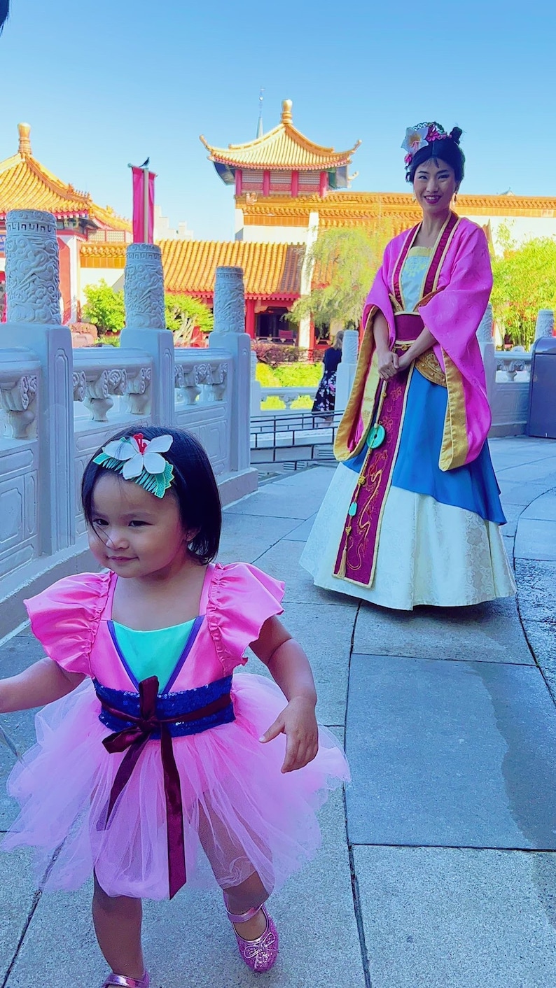 May include: A young girl in a pink and teal dress with a flower in her hair walks towards a woman in a pink and blue dress. The woman is standing in front of a traditional Chinese building.