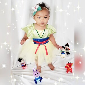 May include: A young girl dressed as Mulan from the Disney movie Mulan. She is wearing a yellow and green dress with a red sash and a blue and silver belt. She is holding a small plush toy of Mulan.