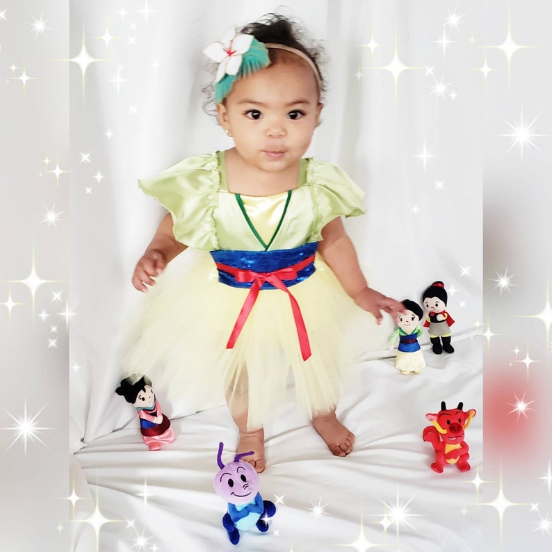 May include: A young girl dressed as Mulan from the Disney movie Mulan. She is wearing a yellow and green dress with a red sash and a blue and silver belt. She is holding a small plush toy of Mulan.
