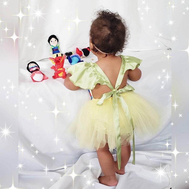May include: A young child wearing a light green satin and yellow tulle dress with a bow in the back. The child is playing with plush toys, including a red dragon, a princess doll, and a cartoon character.