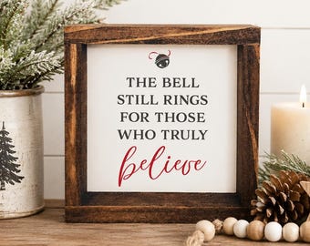 May include: A square wooden sign with a dark brown frame and a white center panel. The text reads THE BELL STILL RINGS FOR THOSE WHO TRULY believe in black and red lettering, with a small graphic of a jingle bell centered above the text.