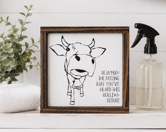 May include: A square wooden sign with a dark brown frame and a white background featuring a black line drawing of a cow wearing a bell. The text reads: DEJA MOO: THE FEELING THAT YOU'VE HEARD THIS BULL$#/+ BEFORE. The sign is displayed on a white tray.