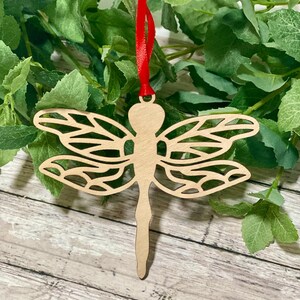 May include: A wooden dragonfly ornament with intricate cutouts, hanging from a red ribbon. The ornament is a light brown color and has a delicate, detailed design.