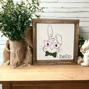 May include: A framed wood sign with a white background featuring a black and white illustration of a bunny wearing pink glasses and a green bow tie. The text "hello" is written in black below the bunny.