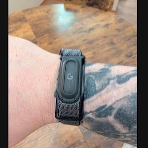 May include: A black fitness tracker with a central oval display featuring a lowercase letter g logo. The device is secured with a textured, dark gray woven fabric strap that wraps around the wrist, providing a secure and adjustable fit.