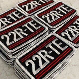 May include: A stack of black and silver embroidered patches with red stitching. The patches feature the text "22R-TE" in white lettering.