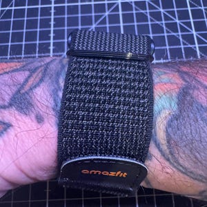 Puede incluir: Una correa de reloj inteligente Amazfit negra y gris se muestra en una muñeca tatuada. La correa está hecha de un material tejido y tiene una hebilla negra. El logotipo de Amazfit es naranja.