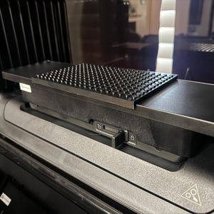 May include: A black rectangular accessory tray with a textured, studded surface designed for a vehicle dashboard. The tray features a central sliding locking mechanism with unlock and lock icons, and a small triangular warning symbol on the base.