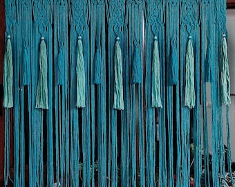 May include: A teal blue macrame wall hanging with fringe tassels. The hanging is made of knotted cords and has a geometric pattern.