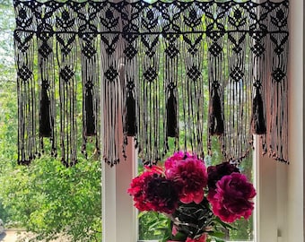 May include: A black macrame curtain with tassels hangs in a window. The curtain is made of thick cord and has a geometric pattern. A bouquet of pink peonies sits on the windowsill.