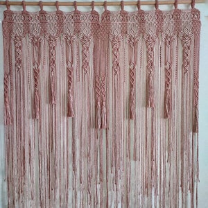 Pode incluir: Uma tapeçaria de macramé de parede em rosa e branco. A tapeçaria apresenta um padrão repetitivo de nós e franjas.