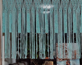 May include: A light blue macrame curtain with tassels hangs in a window. The curtain is made of knotted string and has a decorative pattern. There are string lights above the curtain. There are three decorative items on the windowsill: a wooden chest, a green potted plant, and a small decorative object.