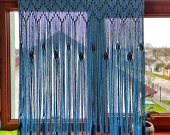 May include: A blue macrame curtain with tassels hanging in a window. The curtain is made of knotted cords and has a diamond pattern.