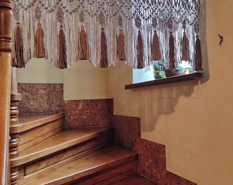 May include: A white and beige macrame wall hanging with tassels hangs above a wooden staircase. The staircase has a cork-like surface on the wall.