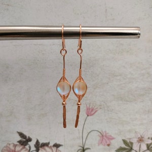 May include: A pair of rose gold earrings with iridescent glass beads and a wire wrapped design. The earrings have a long, thin, hammered wire dangle.