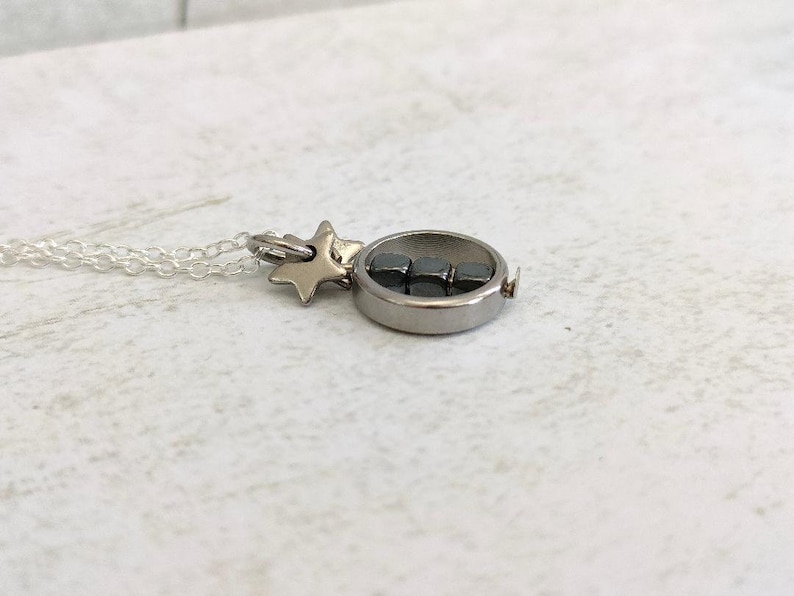 Puede incluir: Un collar de plata con un dije de estrella y un dije redondo que contiene peque&ntilde;os cubos negros.