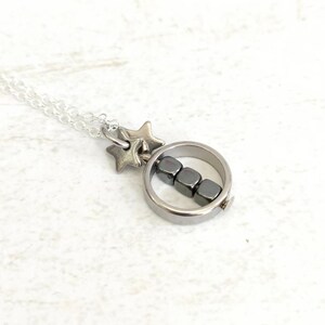 Puede incluir: Un collar de plata con un colgante circular que contiene una fila de cuatro cuentas de hematites negras. El collar tambi&eacute;n tiene un dije de estrella de plata.