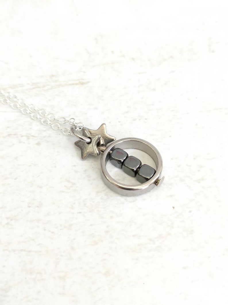 Puede incluir: Un collar de plata con un colgante circular que contiene una fila de cuatro cuentas de hematites negras. El collar tambi&eacute;n tiene un dije de estrella de plata.