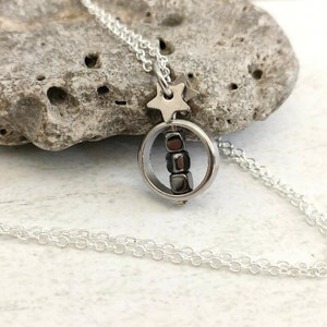 Puede incluir: Un collar de cadena de plata con un peque&ntilde;o colgante redondo que presenta una fila de cuentas negras en su interior. Un dije de estrella plateada cuelga sobre el colgante.