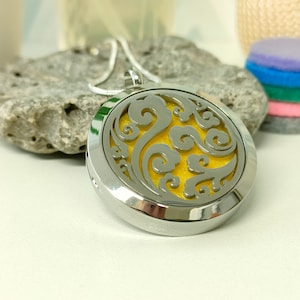 May include: A silver aromatherapy pendant necklace with a swirling design. The pendant has a yellow interior and hangs on a silver chain. A stack of colourful felt pads is visible in the background.