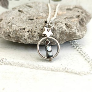 Puede incluir: Un collar de cadena de plata con un dije de estrella y un dije redondo que contiene una pila de peque&ntilde;as cuentas grises oscuras.