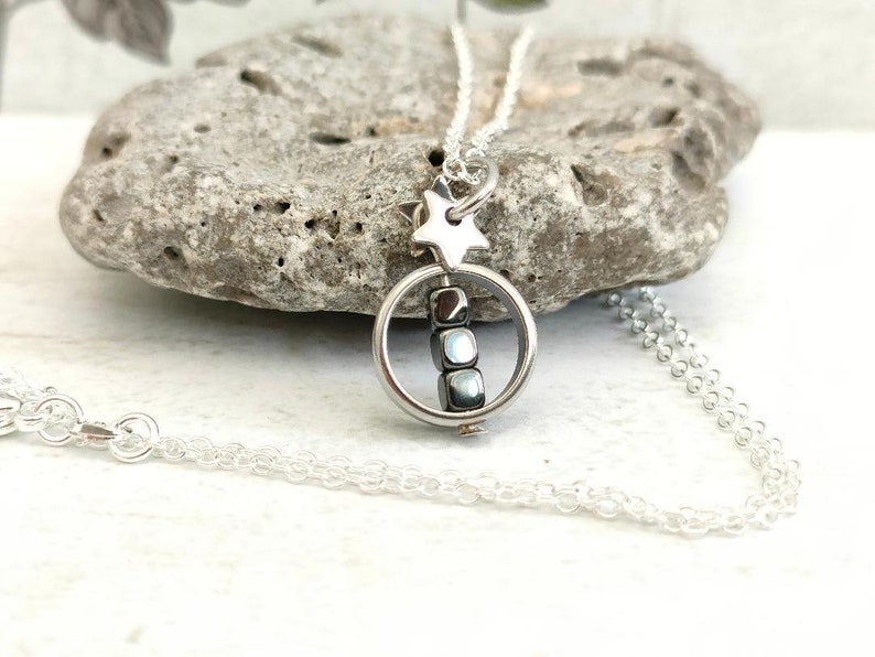 Puede incluir: Un collar de cadena de plata con un dije de estrella y un dije redondo que contiene una pila de peque&ntilde;as cuentas grises oscuras.