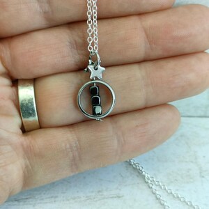 Puede incluir: Un collar de plata con un dije de estrella y un dije circular que contiene cuentas negras. El collar est&aacute; sobre una superficie blanca.