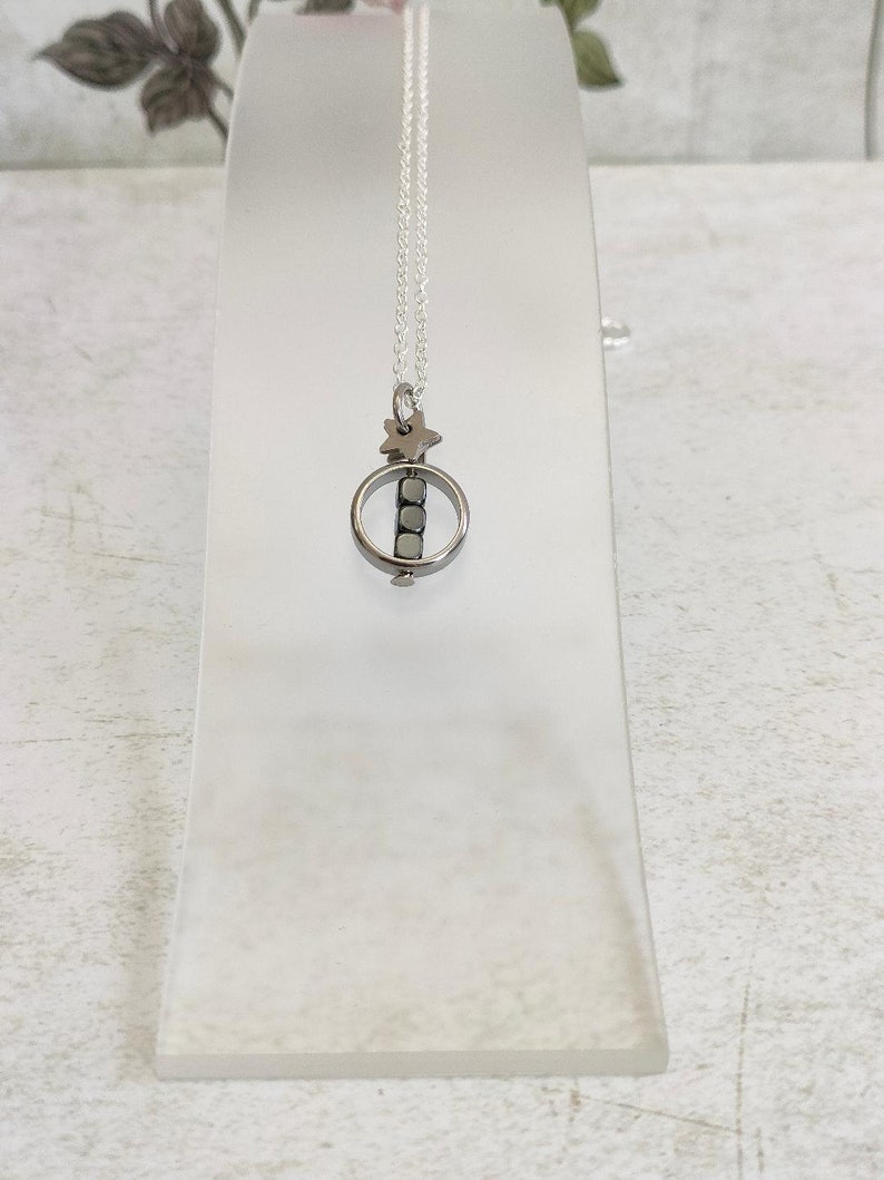 Puede incluir: Un collar de cadena de plata con un peque&ntilde;o colgante que presenta un c&iacute;rculo con una estrella en la parte superior y cuatro cuentas de plata en su interior.