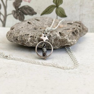 Puede incluir: Un collar de plata con un dije de estrella y un dije redondo que contiene una fila de cuentas negras. El collar est&aacute; sobre una superficie blanca.