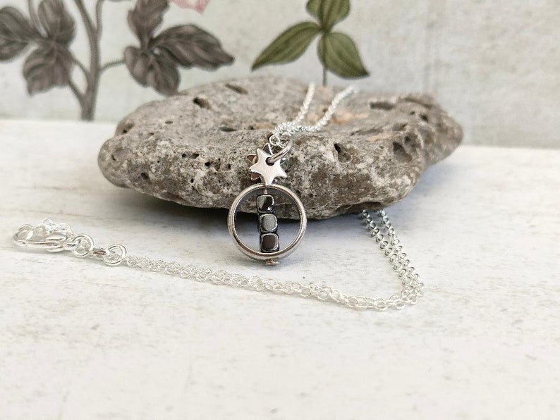 Puede incluir: Un collar de plata con un dije de estrella y un dije redondo que contiene una fila de cuentas negras. El collar est&aacute; sobre una superficie blanca.