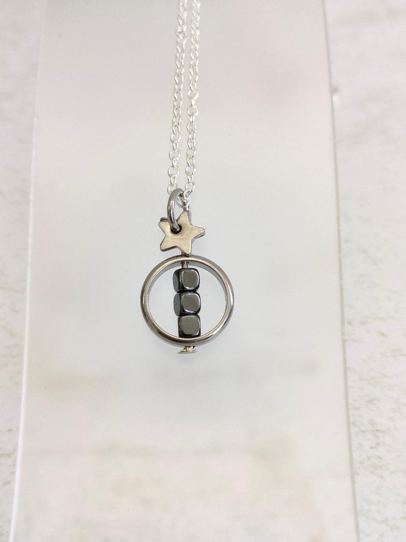 Puede incluir: Un collar de cadena de plata con un dije de estrella de plata y un anillo de plata con cuatro cuentas c&uacute;bicas negras colgando.