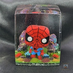 May include: A red and blue Spider-Man figurine with a white background is encased in a clear resin cube. The cube is decorated with green and brown moss and colorful rocks.