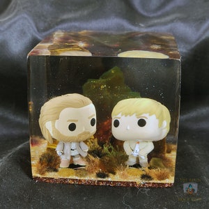 May include: Two small figurines of men with beards are encased in a clear cube filled with a glittery, brown, and green resin. The cube has a brown base.