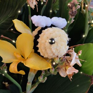 May include: A handmade, plush bee with a yellow body, black stripes, and white wings. The bee is set against a backdrop of yellow flowers and green foliage, creating a whimsical, nature-inspired scene.