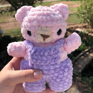Op de afbeelding: Een handgemaakte gehaakte teddybeer in zachtroze en lavendel. De beer heeft zwarte ogen, een bruine snuit en draagt een lavendelkleurige overall. Het pluche speelgoed wordt tegen een vage groene achtergrond gehouden.