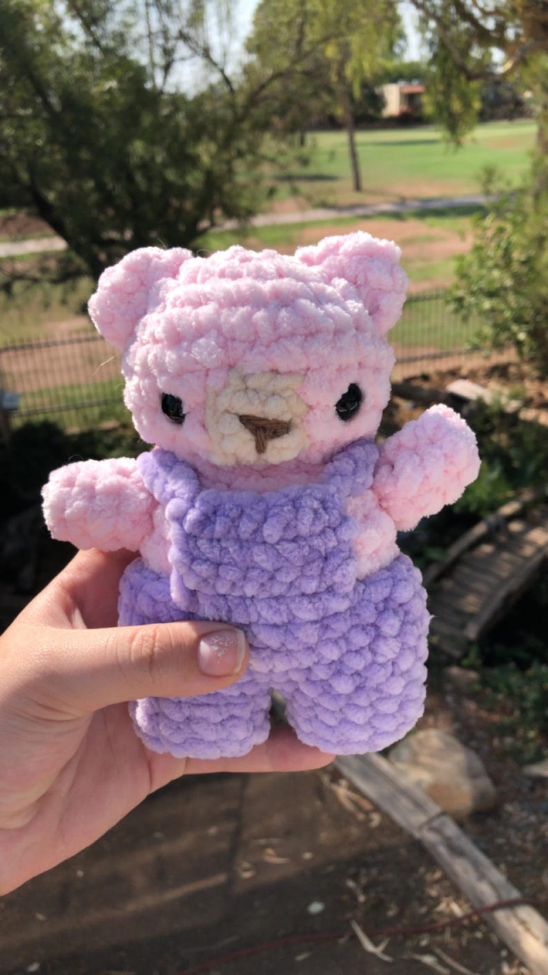 Op de afbeelding: Een handgemaakte gehaakte teddybeer in zachtroze en lavendel. De beer heeft zwarte ogen, een bruine snuit en draagt een lavendelkleurige overall. Het pluche speelgoed wordt tegen een vage groene achtergrond gehouden.