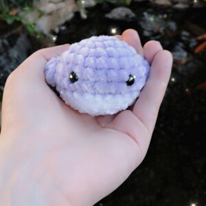 May include: A small, handmade crochet whale in shades of lavender and white. The whale has black safety eyes and a smiling expression. The plush toy is held in a hand, with a blurred background of a pond and greenery.