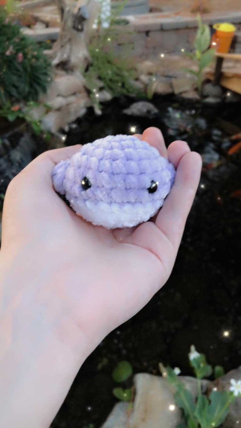 May include: A small, handmade crochet whale in shades of lavender and white. The whale has black safety eyes and a smiling expression. The plush toy is held in a hand, with a blurred background of a pond and greenery.