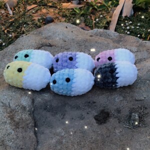 May include: A collection of handmade, plush, oval-shaped toys in various pastel colors, including yellow, blue, purple, and pink, each with a contrasting white section and black bead eyes. The toys are arranged on a natural stone surface.