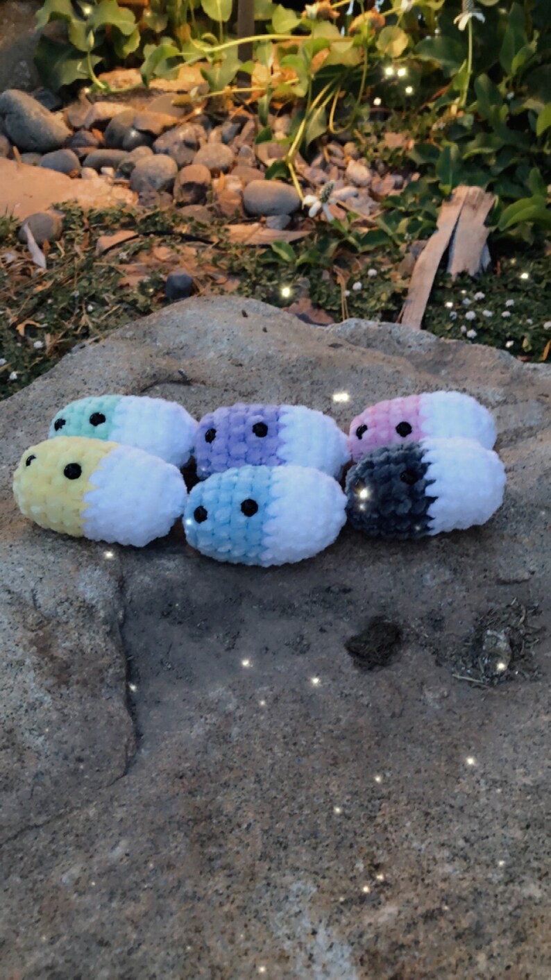 May include: A collection of handmade, plush, oval-shaped toys in various pastel colors, including yellow, blue, purple, and pink, each with a contrasting white section and black bead eyes. The toys are arranged on a natural stone surface.