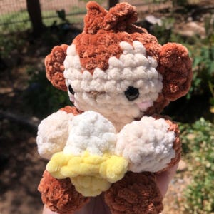 May include: A handmade crochet monkey plushie in shades of brown, white, and yellow. The plush toy has a detailed design with a fluffy texture, black eyes, and a cute expression. The monkey is holding a yellow object.