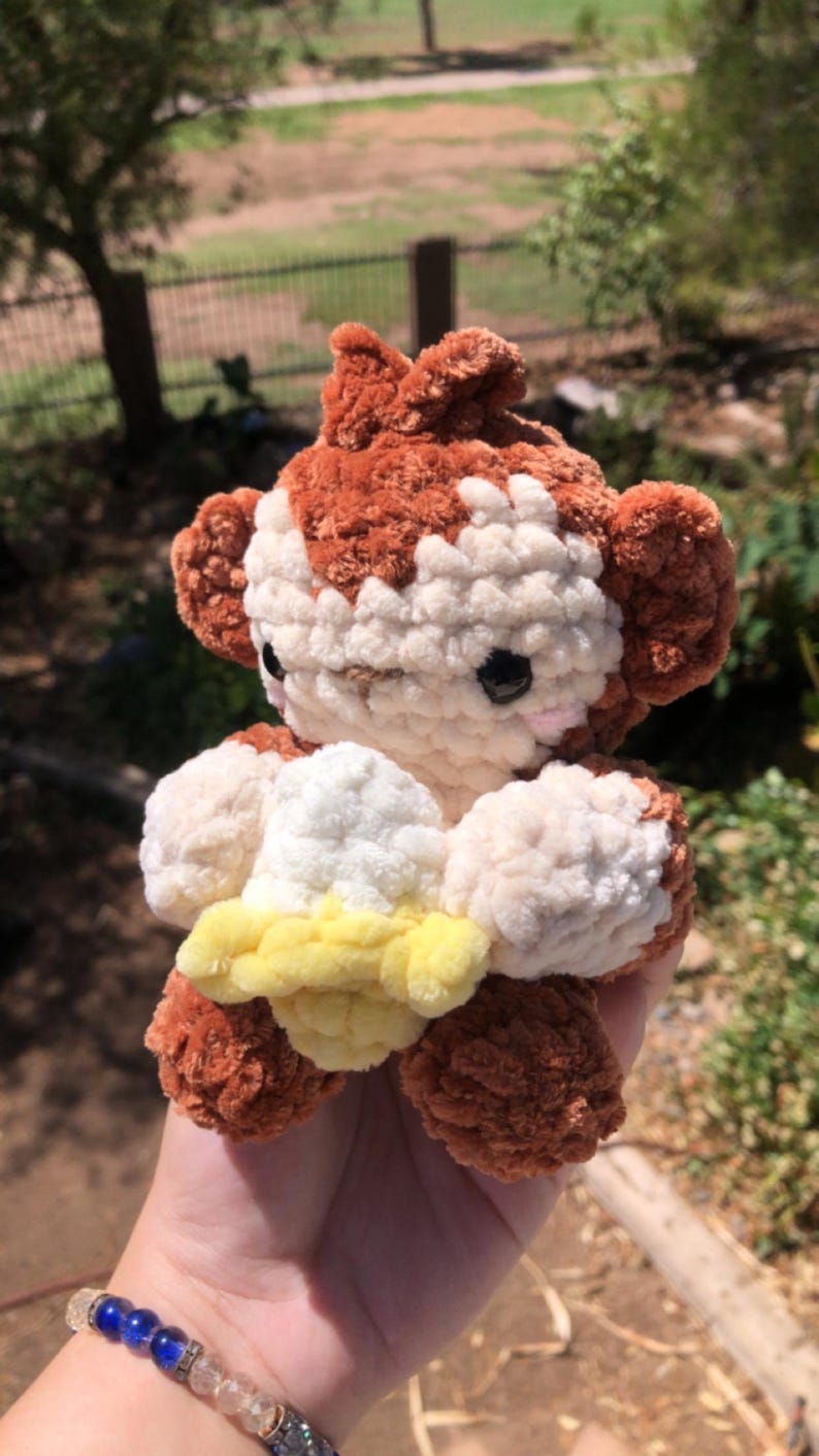May include: A handmade crochet monkey plushie in shades of brown, white, and yellow. The plush toy has a detailed design with a fluffy texture, black eyes, and a cute expression. The monkey is holding a yellow object.